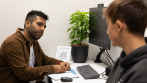 A Health Coach from the BeingWell service supports a young person during a one-to-one session in a clinic room. The Health Coach is taking notes while listening attentively, with a computer and a potted plant on the desk.
