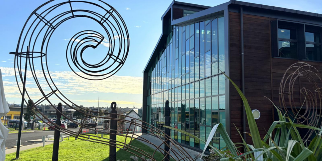 Two-storey modern building with large glass windows and timber cladding at The Kollective in Tauranga, with a large spiral metal sculpture in the foreground and landscaped grass area under a blue sky.