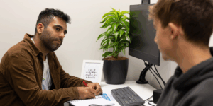 A Health Coach from the BeingWell service supports a young person during a one-to-one session in a clinic room. The Health Coach is taking notes while listening attentively, with a computer and a potted plant on the desk.